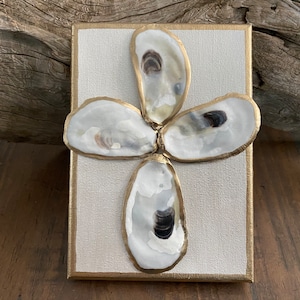 May include: A rectangular canvas art piece featuring four oyster shells arranged in a flower-like pattern. The shells are white with gold-painted edges, set against a cream-colored background with a gold border.