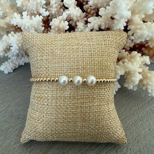 May include: A gold beaded bracelet with three white pearls is displayed on a burlap pillow. The bracelet is set against a backdrop of white coral, creating a contrast of textures and colors. The bracelet is a piece of jewelry.