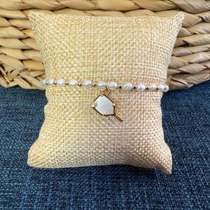 May include: A delicate bracelet featuring white pearls and gold beads. A gold charm in the shape of a tennis racket is suspended from the bracelet. The bracelet is displayed on a small, square, beige pillow.