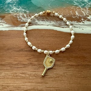 May include: A gold-toned bracelet featuring white pearls and a gold pickleball paddle charm. The paddle charm is embellished with small, clear stones. The bracelet is set against a wooden surface with a blue and white background.