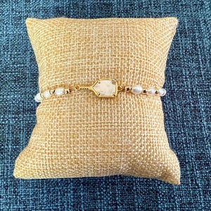 May include: A gold-toned bracelet with white pearls and a central, light-colored, geometric stone. The bracelet is displayed on a small, square, tan burlap pillow against a textured blue background. The jewelry piece is delicate and elegant.