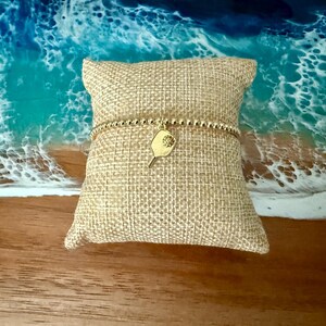 May include: A gold beaded bracelet with a pickleball paddle charm is displayed on a burlap pillow. The charm has a small, textured design. The background features a beach scene with blue and white waves.