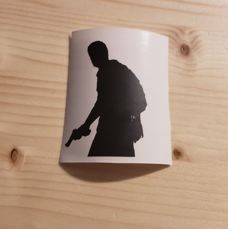 May include: A black silhouette sticker of a person holding a handgun. The figure is facing left, with a backpack visible. The sticker is on a white background, with a wooden surface in the background.