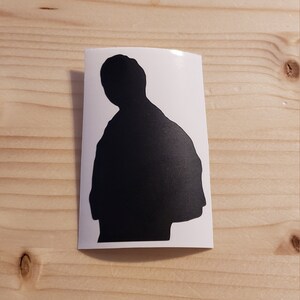 May include: A black silhouette sticker of a person, set against a white background. The sticker is rectangular with rounded corners. The image is on a light-colored wooden surface.