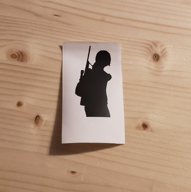 May include: A white sticker featuring a black silhouette of a person carrying a rifle and a backpack. The person is wearing a helmet. The sticker is rectangular and is placed on a light-colored wooden surface.