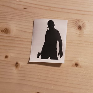 May include: A black silhouette sticker of a person holding a gun. The sticker is on a white background and is placed on a light brown wooden surface.