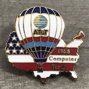 May include: A vintage AT&T Computer Team pin from 1988. The pin features a hot air balloon design with the AT&T logo, set against a map of the United States. The balloon and map incorporate red, white, and blue colors.