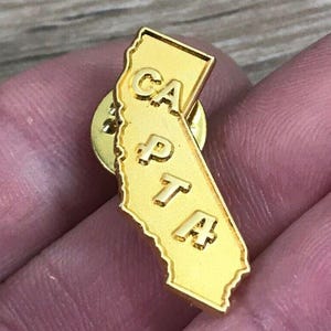 May include: A gold-colored pin in the shape of California, with the letters "CA PTA" embossed on the surface. The pin is a detailed representation of the state's outline.