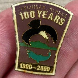 May include: A rectangular pin with a gold border and a maroon background. The pin features the text "Ketchikan, Alaska 100 Years" and the years "1900-2000". The pin has a black and white bird, green mountains, and a teal whale design.