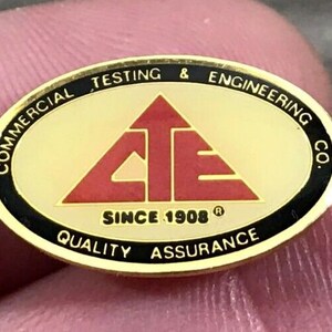 May include: A gold-colored oval pin with black trim. The pin features the text "COMMERCIAL TESTING & ENGINEERING CO." around the edge. Inside is a red triangle with the letters "CTE" and the words "SINCE 1908 QUALITY ASSURANCE".