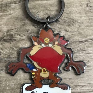 May include: Keychain featuring the Tasmanian Devil character from Looney Tunes, wearing a cowboy hat and holding a gun. The keychain has a silver border and a metal ring. The Six Flags logo is at the bottom.