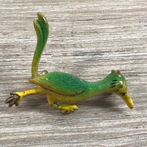 May include: A small, colorful metal figurine of a roadrunner bird. The bird is primarily green and yellow, with a long, curved tail and a yellow beak. The figurine is lying on a light-colored, wood-grain surface.