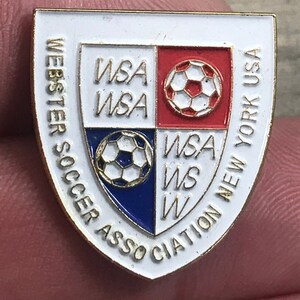 May include: A white, red, and blue enamel pin with a gold border. The pin features a soccer ball and the text "Webster Soccer Association New York USA" and "WSA".