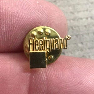 May include: A gold-colored Fleetguard lapel pin. The pin is round with the word "Fleetguard" in a bold, sans-serif font. A small square is below the text. The pin has a classic, vintage design.