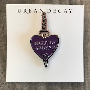 May include: A purple heart-shaped pin with the words "MAKEUP JUNKIE" in white lettering. The pin is designed to look like a dagger, with a silver handle and blade. The pin is on a white card with "URBAN DECAY" printed at the top.