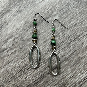 May include: A pair of dangle earrings featuring silver-toned oval pendants. The earrings have a series of green beads and small gold-toned beads. The earrings hang from silver-toned ear wires. The earrings are displayed on a gray wooden surface.