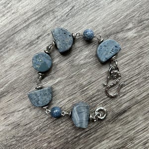 May include: A handmade bracelet featuring blue-grey stone beads and silver-toned chain links. The bracelet has a variety of stone shapes, including half-moon and round, and is secured with a decorative clasp. The bracelet is displayed on a wooden surface.