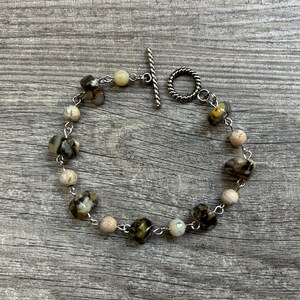 May include: A beaded bracelet featuring a mix of light beige and dark, speckled beads. The bracelet is strung on a silver chain and has a toggle clasp. The beads are irregularly shaped, adding a unique touch to the jewelry.