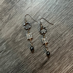 May include: A pair of dangle earrings with silver-tone metal components. Each earring features a textured ring, a square charm, a gold-tone connector, and a dark gray teardrop bead. The earrings hang from silver-tone ear wires.