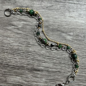 May include: A multi-strand bracelet featuring green and red gemstone beads, gold-tone and silver-tone chains, and a lobster clasp. The bracelet has a layered design with a mix of textures and colors, creating a unique accessory.