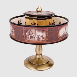 Puede incluir: Un zoótropo vintage con una base dorada y un tambor giratorio. El tambor presenta ilustraciones de figuras en movimiento, con una banda exterior decorativa en burdeos y dorado. La parte superior está adornada con un pináculo dorado.
