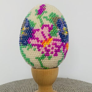 May include: A beaded crochet Easter egg kit with a floral design in shades of pink, purple, blue, green, and white. The egg is displayed on a wooden stand.