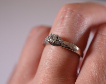 Pode incluir: Um anel de prata com um único diamante redondo e diamantes menores na banda.