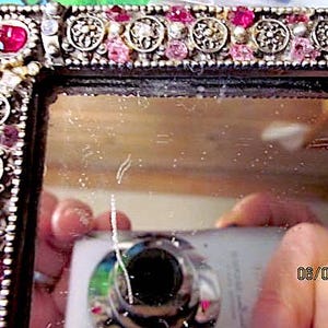May include: Close-up of a decorative mirror with an ornate frame. The frame is silver-toned and embellished with pink and clear gemstones, along with detailed metalwork. The mirror reflects a camera and a hand.