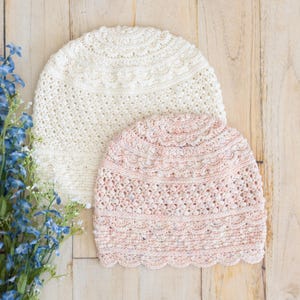 Puede incluir: Dos gorros de ganchillo, uno en color crema sólido y otro en rosa claro moteado, ambos con intrincados patrones circulares en la coronilla y un borde festoneado en el dobladillo inferior, junto a un racimo de pequeñas flores azules.