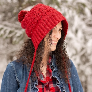May include: A red knitted winter hat with ear flaps and a pom-pom on top. The hat is worn with a denim jacket and a red plaid shirt. The hat's ear flaps have pom-poms at the end.