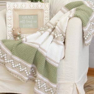 Op de afbeelding: Een handgemaakte gehaakte deken in wit, groen en beige, gedrapeerd over een crèmekleurige fauteuil. De deken heeft getextureerde steken en decoratieve bobbels. Een ingelijst bord met de tekst "GRACE will lead me HOME" is zichtbaar.