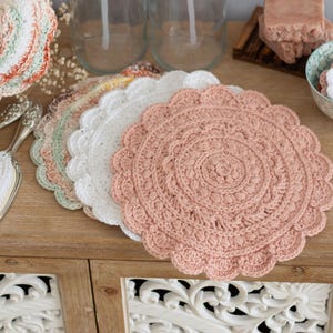Könnte beinhalten: Eine Sammlung handgefertigter Häkelarbeiten in Pastellfarben. Das Bild zeigt mehrere runde Deckchen mit gewelltem Rand in Rosa, Weiß und Grün. Ein Vintage-Spiegel und eine Bürste sind ebenfalls zu sehen, was auf ein Badezimmer- oder Schminktisch-Thema hindeutet.