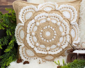 May include: A square throw pillow featuring a tan crocheted base with an intricate, raised white floral mandala pattern centered on the front. The design includes textured bobble stitches and scalloped edges, creating a layered, dimensional look.