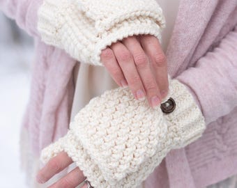 May include: Cream-colored, crocheted fingerless gloves with a button closure. The gloves are made of a thick yarn and have a textured stitch pattern. A brown button secures the cuff. The gloves are shown against a soft pink cardigan.