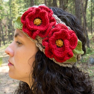 Pode incluir: Uma faixa de cabeça de crochê com duas flores vermelhas e folhas verdes. A faixa de cabeça é feita de fio marrom.