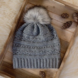 May include: A gray crocheted beanie with a white faux fur pom pom. The beanie is in a wooden tray with pine cones.