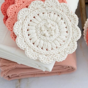 May include: A collection of handmade crochet coasters in shades of white, peach, and coral. The coasters have a scalloped edge and a floral center design. They are stacked on folded white and peach towels.