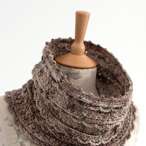 Puede incluir: Un cuello de ganchillo marrón y beige con un diseño texturizado y en capas. El cuello se exhibe en un maniquí y presenta un hilo moteado con motas de color. Un accesorio acogedor.