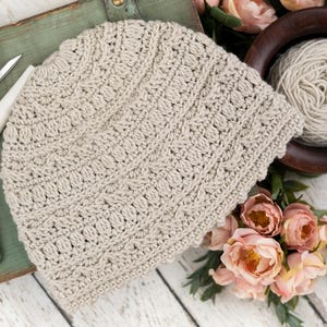 Può includere: Un berretto all'uncinetto beige caratterizzato da un motivo testurizzato con file alternate di punti nocciolina e punti alti. Il cappello è realizzato in filato di colore chiaro e presenta un motivo a punti coerente e ripetitivo.