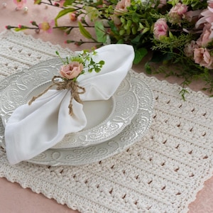 May include: A cream-coloured, crocheted placemat with a scalloped edge, paired with two white, patterned plates. A white napkin is adorned with a small flower arrangement and twine. Floral arrangement in the background.