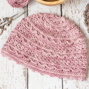 Puede incluir: Un gorro de punto de color rosa con un patrón de puntadas texturizadas. El gorro está colocado plano sobre una superficie de madera blanca.