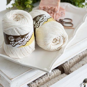 White dresser with crochet dishcloths in a drawer, yarn, soap, scissors, and crochet hook  with flowers on the top.
