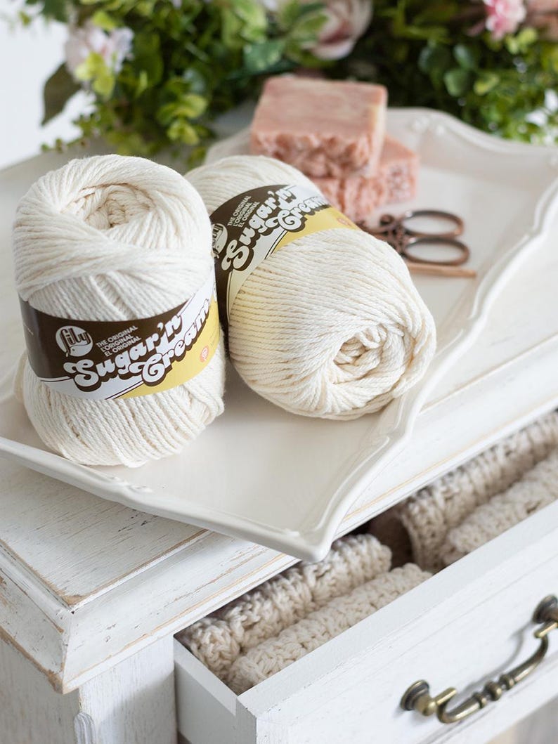 White dresser with crochet dishcloths in a drawer, yarn, soap, scissors, and crochet hook  with flowers on the top.