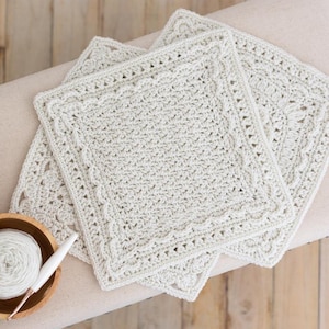 Puede incluir: Tres tapetes cuadrados de ganchillo de color crema con un borde festoneado están dispuestos sobre una superficie neutra. Un cuenco de madera con hilo blanco y un ganchillo se encuentran en la esquina inferior izquierda. Los tapetes tienen un patrón de puntada texturizada.