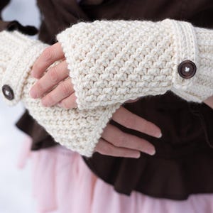 Puede incluir: Guantes sin dedos de crochet color crema con cierre de botón. Los guantes están hechos de un hilo grueso y tienen un patrón de puntada texturizado. Los botones son de color marrón oscuro. Los guantes se muestran sobre un fondo borroso.