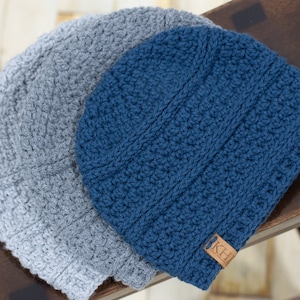 May include: Two crocheted hats, one gray and one blue. The blue hat has a brown leather tag with the letters "KH" on it.