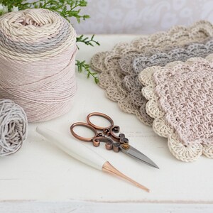 Pile of crochet scalloped mug rugs with yarn, scissors, flowers, and crochet hook.