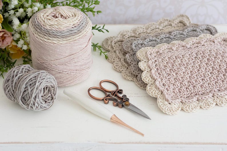 Pile of crochet scalloped mug rugs with yarn, scissors, flowers, and crochet hook.
