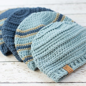 May include: Four crocheted hats in shades of blue and beige. The hats are stacked on top of each other, with the largest hat on the bottom and the smallest hat on top. The hats are all made with a simple stitch pattern and have a textured look. The hat on top has a brown leather tag with the word "KHT" on it.