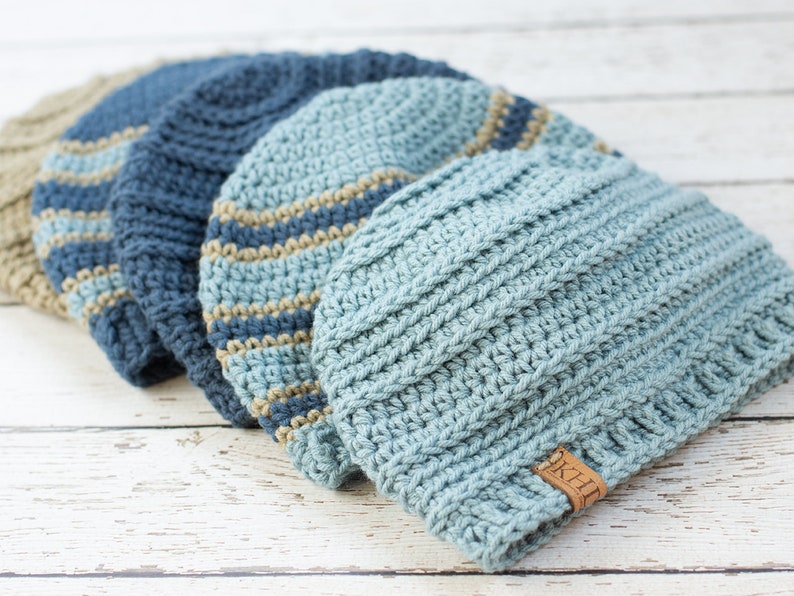 May include: Four crocheted hats in shades of blue and beige. The hats are stacked on top of each other, with the largest hat on the bottom and the smallest hat on top. The hats are all made with a simple stitch pattern and have a textured look. The hat on top has a brown leather tag with the word "KHT" on it.